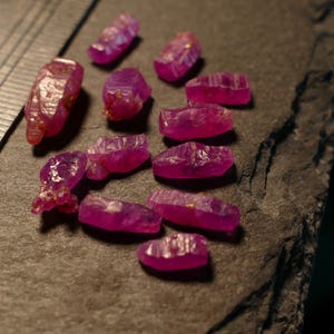 May include: A collection of eleven small, raw, vibrant pinkish-red rubies with irregular, faceted-like surfaces. A metal ruler is positioned on the left side, showing the stones range from approximately 0.1 to 0.5 inches in length.
