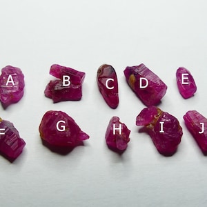 Könnte beinhalten: Eine Sammlung von zehn grob geschnittenen, tiefroten Rubin-Edelsteinen, jede mit einer einzigartigen Form und Textur. Die Edelsteine sind mit A bis J beschriftet und werden vor einem schlichten weißen Hintergrund präsentiert. Die Rubine variieren in der Größe und haben ein natürliches, unpoliertes Aussehen.