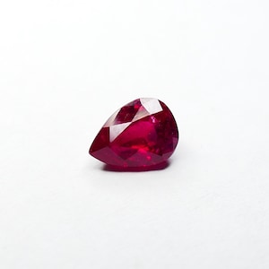 May include: A faceted, pear-shaped ruby gemstone. The gem is a deep, saturated red colour, with multiple facets catching the light. The ruby is set against a plain white background, highlighting its brilliance.