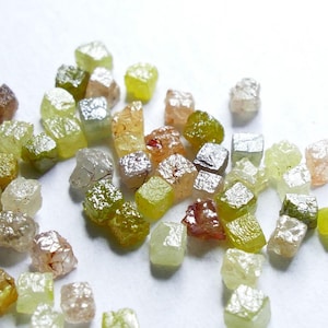 Peut inclure: Un ensemble de diamants bruts non taillés de différentes couleurs, dont le jaune, le vert, le marron et le transparent. Les pierres précieuses sont dispersées sur une surface blanche, révélant leurs formes et facettes naturelles et irrégulières.