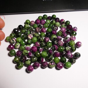 Pode incluir: Uma coleção de contas de pedras preciosas facetadas em tons de verde, roxo e preto. As contas são redondas e têm cerca de 0,6 cm de diâmetro. Estão espalhadas numa superfície branca.