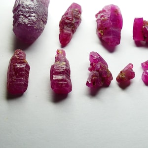 May include: A collection of raw, uncut ruby crystals in varying sizes and shapes. The crystals display a deep, vibrant pinkish-red hue, with rough, textured surfaces. The gemstones are arranged on a white surface, showcasing their natural beauty.