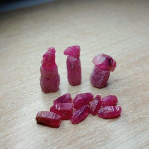 May include: A collection of raw, uncut ruby gemstones in varying shapes and sizes. The rubies display a deep, rich red color and are scattered on a light-colored wooden surface. Some stones are elongated, while others are more compact.