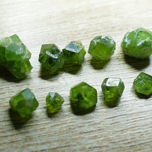 Könnte beinhalten: Eine Sammlung leuchtend grüner Peridot-Edelsteine, in verschiedenen Größen und Formen, liegt auf einer hellen Holzoberfläche. Die Edelsteine weisen eine kristalline Struktur und eine durchscheinende Qualität auf.