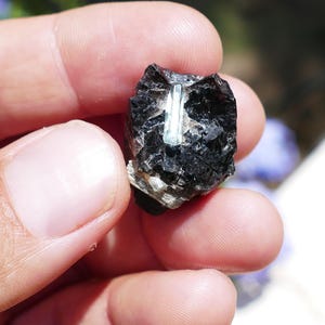 Könnte beinhalten: Ein dunkles, rau strukturiertes Mineralexemplar mit einem hellblauen Kristalleinschluss. Das Mineral wird zwischen den Fingern gehalten, was sein natürliches, unpoliertes Aussehen zeigt. Der Hintergrund ist unscharf, wodurch die Details des Steins hervorgehoben werden.