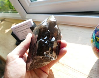 May include: A dark brown, faceted crystal with a pointed top is held in a hand. The crystal has a glossy, reflective surface. A colorful glass globe and a business card holder are in the background.