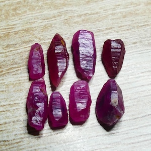 Może przedstawiać: Kolekcja dziewięciu surowych kamieni szlachetnych rubinu. Kamienie, o różnych rozmiarach i kształtach, prezentują głęboki czerwono-fioletowy odcień z widocznymi strukturami krystalicznymi. Ułożone na jasnej drewnianej powierzchni.