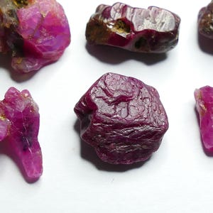 Puede incluir: Colección de gemas de rubí en bruto de diversas formas y tamaños. Las gemas muestran un color rosa y rojo intenso, con superficies rugosas y texturizadas. La disposición muestra la belleza natural y sin pulir de los rubíes.