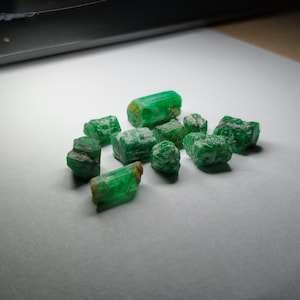 Pode incluir: Uma coleção de pequenos cristais de esmeralda brutos em vários tons de verde. As pedras apresentam texturas naturais não polidas com formas geométricas irregulares e superfícies translúcidas a opacas, espalhadas sobre uma superfície de cor clara.