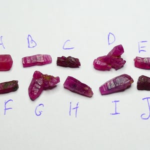 Puede incluir: Diez cristales de rubí en bruto, de color rosa intenso a rojo, dispuestos en dos filas y etiquetados de la A a la J con tinta azul manuscrita. Cada piedra muestra una estructura cristalina única, irregular y con texturas naturales.
