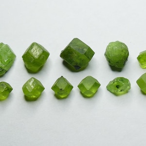 May include: A collection of eleven faceted, bright green peridot gemstones of varying sizes. The gemstones are arranged on a white surface, showcasing their natural, crystalline structures and vibrant color.