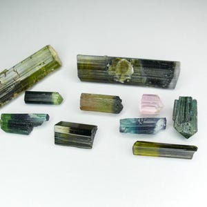 以下が含まれることがあります： 緑、黒、ピンク、青など、さまざまな色の生のトルマリン結晶の詰め合わせ。 結晶は白い表面に表示され、自然な質感と形状が示されています。 コレクターやジュエリー作りに最適です。