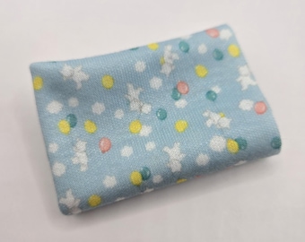 Pode incluir: Uma peça de tecido azul-claro dobrada com um padrão repetitivo de silhuetas de ursos de peluche brancos, círculos amarelos, pequenos pontos azul-petróleo e formas circulares rosa-suave espalhadas pela superfície em um arranjo denso.