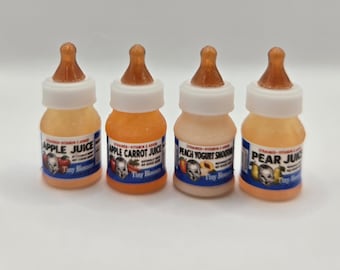 Pode incluir: Quatro biberões em miniatura com tampas brancas e tetinas cor de âmbar. Os rótulos dizem Apple Juice, Apple Carrot Juice, Peach Yogurt Smoothie e Pear Juice, cada um com uma ilustração de um rosto de bebé e a marca Tiny Blossom.
