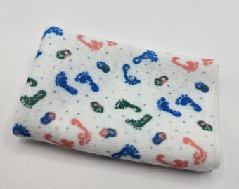 Pode incluir: Um pano de tecido retangular branco com um padrão repetitivo de pequenas pegadas nas cores azul, verde e coral. O design é intercalado com pequenos pontos multicoloridos e pequenos ícones quadrados contendo a letra A.