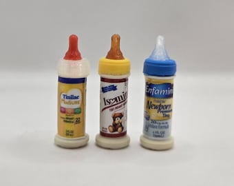 Pode incluir: Três biberões em miniatura com rótulos que dizem Tinilac, Isomin e Enfamil. Cada biberão apresenta uma tampa e tetina coloridas, com texto indicando uma capacidade de 59 ml. Os biberões estão dispostos em fila contra um fundo liso.
