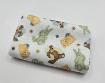 Pode incluir: Uma peça de tecido branco dobrada com um padrão repetitivo de pequenos ursos castanhos e coelhos verde-sálvia ilustrados. Os ursos estão representados em várias posições, alguns parecendo dormir ou descansar, espalhados pelo fundo branco.