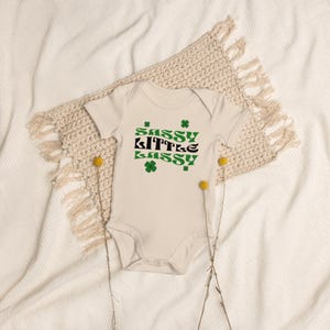 May include: A cream-colored baby onesie with short sleeves and a green and black graphic that says "Sassy Little Sassy" with shamrock designs.