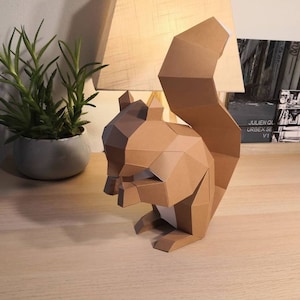 Op de afbeelding: Een bruine papieren eekhoorn sculptuur met een pluche staart. De eekhoorn zit op een houten oppervlak met een lamp op de achtergrond.