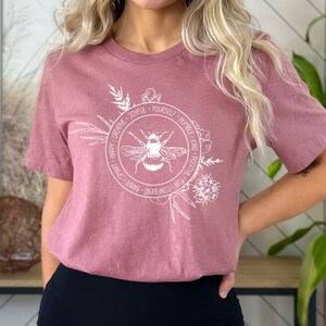 May include: Dusty rose-colored t-shirt featuring a white circular design with a bee illustration. The circle is surrounded by floral elements and words like "Happy", "Creative", and "Joyful". The shirt has short sleeves and a crew neck.