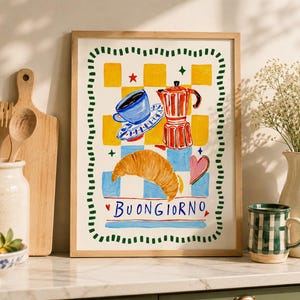 May include: A framed art print featuring a colorful illustration of a blue coffee cup, a red moka pot, and a golden croissant on a yellow and blue checkered background. The text BUONGIORNO is written at the bottom, surrounded by a green dashed border.