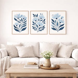 Puede incluir: Tres impresiones art&iacute;sticas florales enmarcadas. Cada impresi&oacute;n presenta una disposici&oacute;n estilizada de flores azules y hojas azul oscuro sobre un fondo blanco. Los marcos son de color madera clara. Las impresiones se exhiben sobre un sof&aacute; blanco.