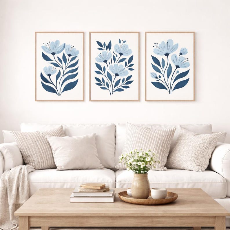 Puede incluir: Tres impresiones art&iacute;sticas florales enmarcadas. Cada impresi&oacute;n presenta una disposici&oacute;n estilizada de flores azules y hojas azul oscuro sobre un fondo blanco. Los marcos son de color madera clara. Las impresiones se exhiben sobre un sof&aacute; blanco.