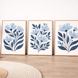 Puede incluir: Tres impresiones art&iacute;sticas florales enmarcadas. Cada impresi&oacute;n presenta un dise&ntilde;o estilizado de flores azules y hojas azul oscuro sobre un fondo crema. Los marcos son de color madera clara. Las impresiones se exhiben sobre un suelo de madera.