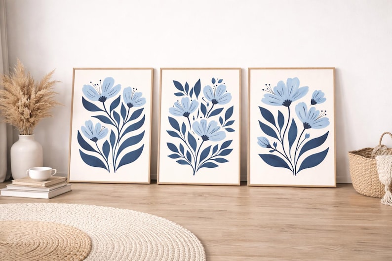 Puede incluir: Tres impresiones art&iacute;sticas florales enmarcadas. Cada impresi&oacute;n presenta un dise&ntilde;o estilizado de flores azules y hojas azul oscuro sobre un fondo crema. Los marcos son de color madera clara. Las impresiones se exhiben sobre un suelo de madera.
