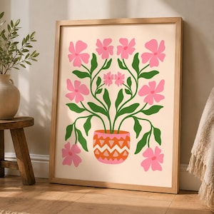 May include: A framed wall art print featuring a symmetrical illustration of pink flowers with green stems and leaves growing out of a patterned orange and pink pot. The artwork is set against a light cream background within a light wood frame.