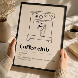 Op de afbeelding: Een ingelijste print met een zwarte rand toont een zwart-wit illustratie van een koffiemachine en een persoon die een koffiekopje vasthoudt. De tekst "Coffee club" wordt eronder weergegeven, samen met een definitie.