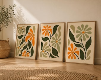 Op de afbeelding: Drie rechthoekige ingelijste wandkunstprints met gestileerde bloemenillustraties. Elke print toont een crèmekleurige achtergrond met gedurfde, effen botanische vormen in tinten van gebrand oranje, saliegroen en donker bosgroen.