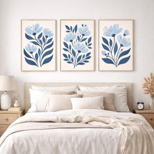 Puede incluir: Tres impresiones art&iacute;sticas florales enmarcadas. Cada impresi&oacute;n presenta un dise&ntilde;o de flor azul estilizada con hojas azul oscuro sobre un fondo crema. Los marcos son de color madera clara. Las impresiones se exhiben sobre una cama con ropa de cama blanca.