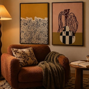 May include: A rust-colored textured armchair with a patterned throw pillow and a draped blanket. Beside it, a floor lamp with a pleated shade, two framed abstract wall art prints, and a round wooden side table holding an open book and a mug.