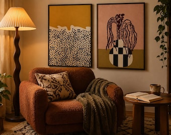 May include: A rust-colored textured armchair with a patterned throw pillow and a draped blanket. Beside it, a floor lamp with a pleated shade, two framed abstract wall art prints, and a round wooden side table holding an open book and a mug.