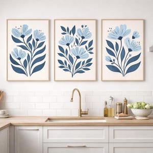 Puede incluir: Tres impresiones art&iacute;sticas florales enmarcadas. Cada impresi&oacute;n presenta un dise&ntilde;o de flores azul claro con hojas y tallos azul oscuro sobre un fondo crema. Los marcos son de color madera clara. Las impresiones se exhiben en una cocina.
