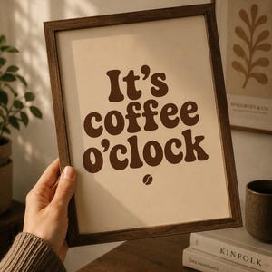 Puede incluir: Una lámina decorativa rectangular en un marco de madera marrón oscuro con el texto It's coffee o'clock en una fuente redondeada, gruesa y de color marrón oscuro sobre un fondo beige claro, con un pequeño icono de grano de café debajo.