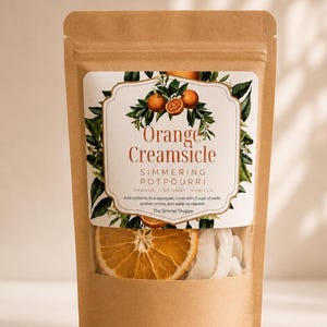 May include: A brown kraft paper pouch containing simmering potpourri. The label features an illustration of oranges and leaves with the text Orange Creamsicle, Simmering Potpourri, Orange, Coconut, Vanilla, and instructions to add 2 cups of water.