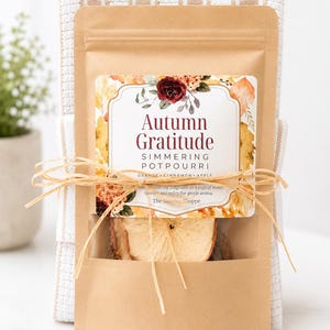 May include: A brown paper pouch of Autumn Gratitude simmering potpourri featuring dried apple slices, tied with raffia string, and placed in front of a white and tan striped waffle-weave kitchen towel. The label displays floral graphics.