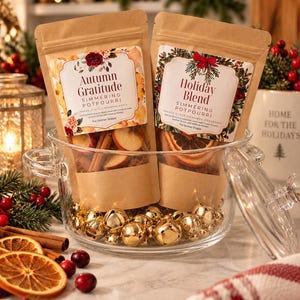 May include: Two brown paper pouches of simmering potpourri labeled Autumn Gratitude and Holiday Blend, placed inside a clear glass bowl filled with gold jingle bells. The labels feature floral and greenery illustrations.