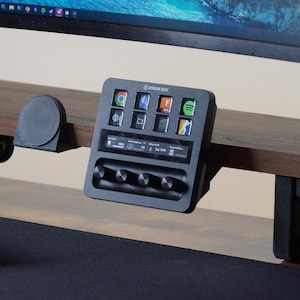 Może przedstawiać: Czarne urządzenie Stream Deck z wieloma ikonami aplikacji wyświetlanymi na drewnianym biurku. Urządzenie posiada kilka pokręteł i przycisków. Monitor komputera i klawiatura są również widoczne w tle.