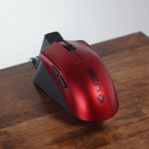 May include: A red computer mouse with the word "SEN" printed on it, resting on a black charging dock. The mouse has a sleek design with buttons and a scroll wheel. The setup is on a wooden surface.