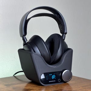 May include: Black over-ear headphones resting in a dark gray charging stand. The stand has a control panel with a dial and the "steelseries" logo. The headphones are on a wooden surface.