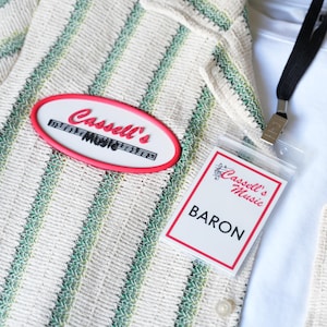 May include: A cream-colored shirt with vertical green and white stripes, featuring an oval red-bordered patch that reads Cassell's Music. A white ID badge with a red border and the text Cassell's Music BARON hangs from a black lanyard.