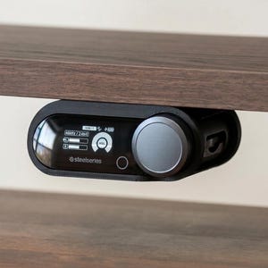 May include: A black SteelSeries audio device with a silver dial and a display screen, mounted under a wooden shelf. The screen displays icons and text. The device is for audio control.