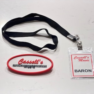 Peut inclure: Un cordon en tissu noir avec un porte-badge en plastique transparent contenant une carte blanche avec le texte rouge Cassell's Music et le texte noir BARON. À côté se trouve un écusson en caoutchouc blanc ovale avec une bordure rouge et le texte Cassell's Music.