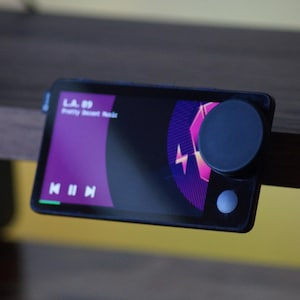 May include: A black rectangular device with a purple screen displaying a music player interface. The screen shows the text "L.A. 69" and "Pretty Sweet Music". The device has a round black button on the right side.