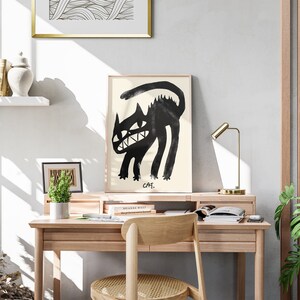 May include: A black and white illustration of a cat with a toothy grin, framed and hanging on a wall above a wooden desk with a lamp, books, and a potted plant.