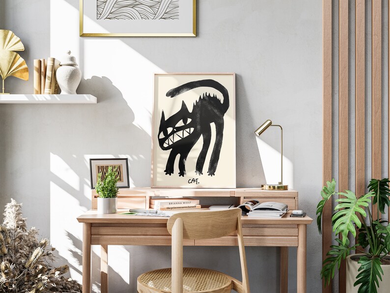 May include: A black and white illustration of a cat with a toothy grin, framed and hanging on a wall above a wooden desk with a lamp, books, and a potted plant.