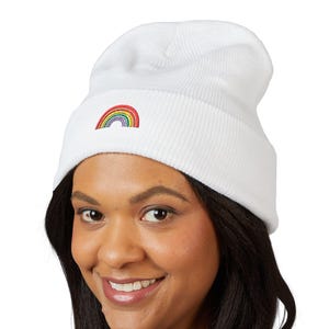 May include: A white ribbed knit beanie featuring a small, embroidered rainbow graphic centred on the front cuff. The hat is constructed with a folded brim design and a soft, textured fabric finish, providing a clean and simple aesthetic.